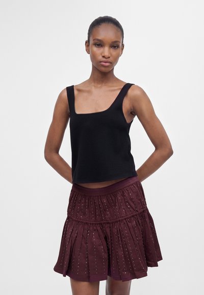 Femme portant un top noir sans manches à encolure carrée et une jupe courte bordeaux avec de petites décorations brillantes, debout les mains derrière le dos.
