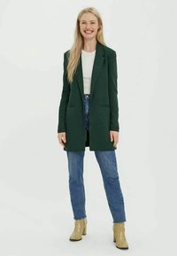 Donna sorridente con lunghi capelli biondi, che indossa un blazer verde scuro, una maglietta bianca, jeans blu e stivaletti beige con tacco, in piedi su uno sfondo bianco.