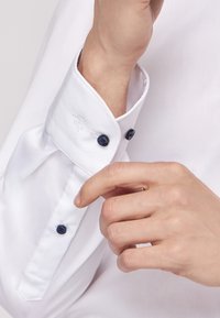 Witte dress shirt met een gesloten manchet. Voorzien van donkerblauwe knopen en subtiele borduurwerk nabij het manchet. Glad, strak textiel.