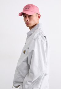 Lys grå jakke med lynlås og høj krave, prydet med et logo patch, kombineret med en pink kasket med sort tekst, båret i en let vinkel.