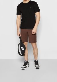 Muž v černém tričku, hnědých šortkách a černo-bílých teniskách, držící černou ledvinku Nike za popruh na jednobarevném pozadí.