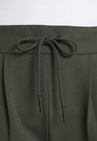 Pantalons taille olive en vert avec une fermeture à cordon, présentant une texture lisse et des plis à l'avant pour un look élégant.