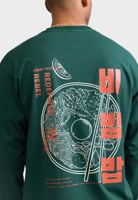 Persona che indossa una maglietta verde scuro con una grande grafica di una ciotola di bibimbap, bacchette e testo in inglese e coreano sul retro.