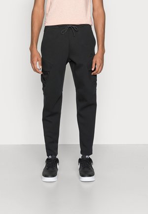 Schwarze Jogginghose aus weichem Stoff mit einem verstellbaren Bund, seitlichen Taschen und schmal zulaufenden Knöcheln, kombiniert mit schwarzen Sneakers.