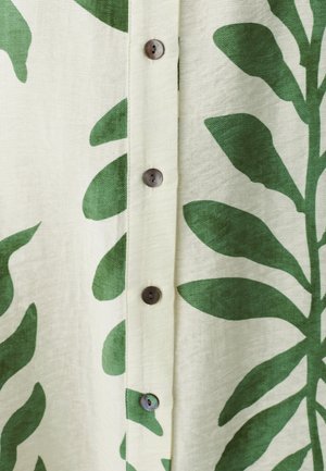 Tissu à motif de feuilles vertes avec un fond beige, comportant des boutons ronds marron le long de la patte de boutonnage centrale. Matériau léger et texturé.