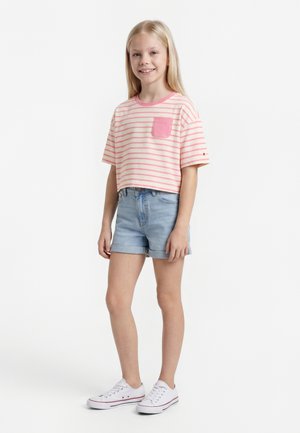Ung pige står og smiler, iført en lyserød og hvid stribet cropped skjorte, lyse denimshorts og hvide sneakers.