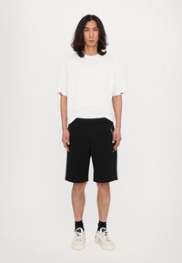 Pantaloni corti neri con tasche laterali, abbinati a una t-shirt bianca a girocollo. Il modello indossa scarpe da ginnastica bianche con calzini neri. Design semplice.
