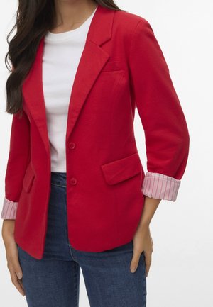 Mujer con blazer rojo brillante con puños de rayas enrollados sobre una camisa blanca y jeans azules, de pie contra un fondo blanco.