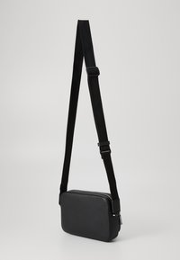 Black rectangular crossbody bag with adjustable nylon strap and silver zipper on a plain light background.