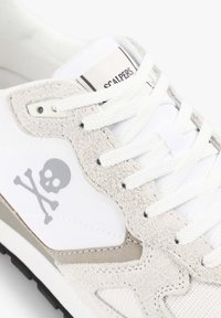 Zapatilla blanca que presenta un panel lateral de ante gris texturizado, un gráfico de calavera en gris y cordones blancos. Incluye una etiqueta con el nombre de la marca.