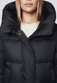 Czarna kurtka puchowa z wysokim kołnierzem, zapięciem na zamek błyskawiczny i dużymi pikowanymi elementami. Gładkie matowe wykończenie z dodatkiem napów.