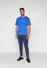 Blå bomulds T-shirt med et hvidt "PARIS 5G" tryk, parret med mørkeblå tapered bukser og sorte atletiske sko.