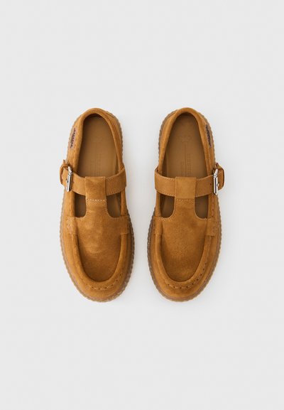 Chaussures plates en daim marron avec un design à barre en T, bout arrondi et semelle en caoutchouc. Elles présentent une boucle argentée et des coutures visibles le long des bords.