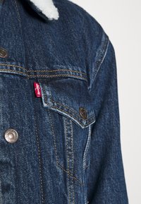 Veste en denim bleu foncé avec boutons-pression métalliques, rabat de poche poitrine et doublure de col en polaire blanche.