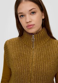 Strickpullover-Weste in Senfgelb mit hohem Kragen, ausgestattet mit einem Reißverschluss vorne mit runder Ziehhilfe, texturiertem Rippstrick und glatten Ärmeln.