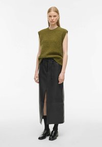 Kaki pletena brezrokavnik v kombinaciji z dolgim črnim usnjenim krilom z razporkom spredaj in pasom na vrvico. Črne sijajne čevlje dopolnjujejo videz.