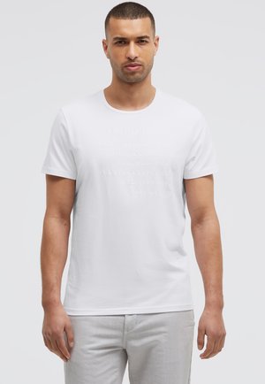 Mann mit kurzen Haaren und leichtem Bart, der ein weißes T-Shirt mit erhabenem Text und hellgrauen Hosen trägt, steht vor einem schlichten Hintergrund.