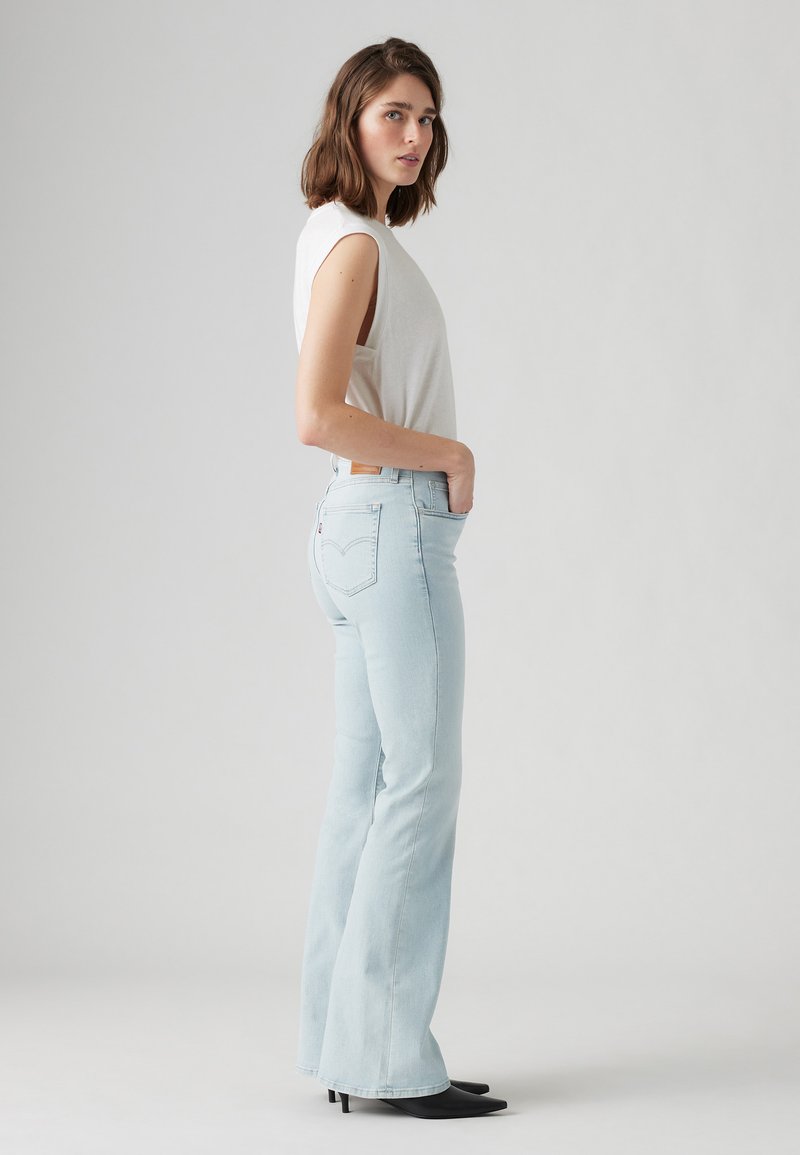 Jeans de mezclilla de tiro acampanado en azul claro, que presentan un diseño clásico de cinco bolsillos y una etiqueta de cuero en la parte posterior. Combinados con una blusa sin mangas blanca.