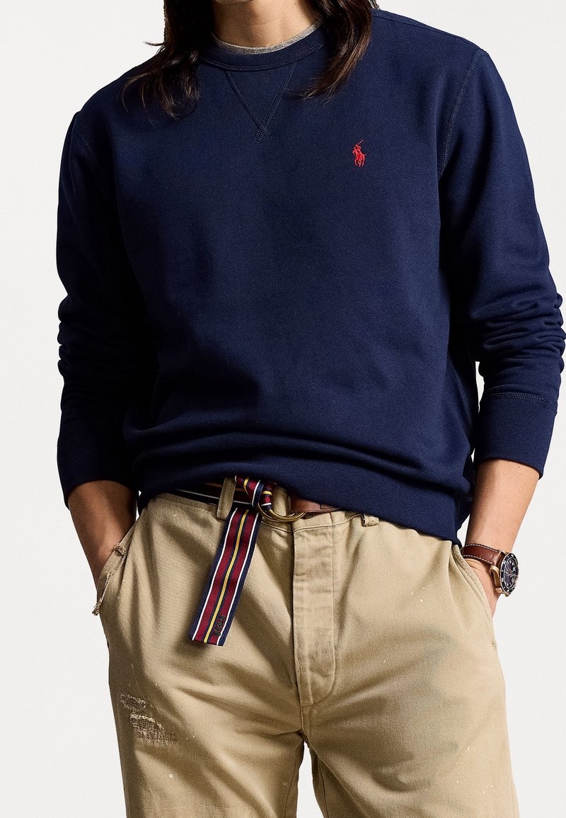 Marinblå sweatshirt med ribbade ärmslut och halsringning, prydd med en liten röd logotyp på bröstet, tillsammans med ljusa, något slitna byxor och ett randigt bälte.