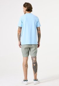 Lichtblauwe katoenen T-shirt, korte mouwen, ronde hals, gecombineerd met getextureerde groene shorts. Zichtbare tatoeages op de benen, witte sneakers.