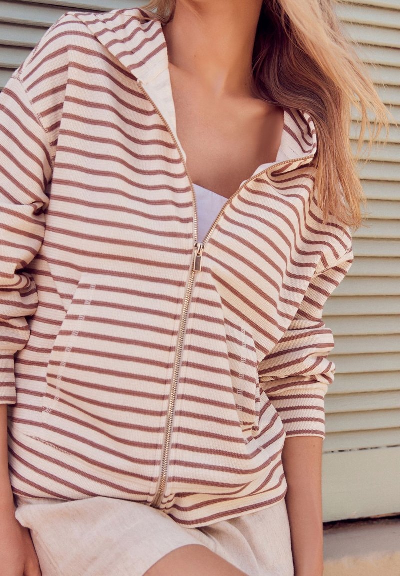 Femme portant un sweat à capuche zippé rayé beige et marron avec manches retroussées, jupe beige, debout devant un volet beige.