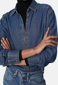 Ljusblå denimskjorta med knapplås framtill, krage och långa ärmar. Har en klassisk passform och synliga sömdetaljer.