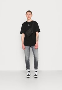 T-shirt oversized preta com logotipo branco, jeans cinzentos, pouch preto e tênis preto e branco; estilo moderno e casual.