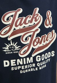 Camiseta azul marina con un gran texto gráfico en blanco y burdeos que dice "Jack & Jones" y "DENIM GOODS". Presenta un logotipo de sol y el detalle "desde 1990".