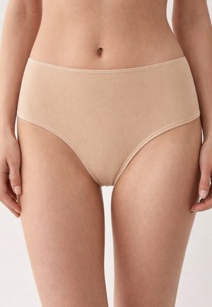 Donna che indossa biancheria intima in cotone beige a vita alta, in piedi con le mani rilassate ai lati, su uno sfondo bianco.