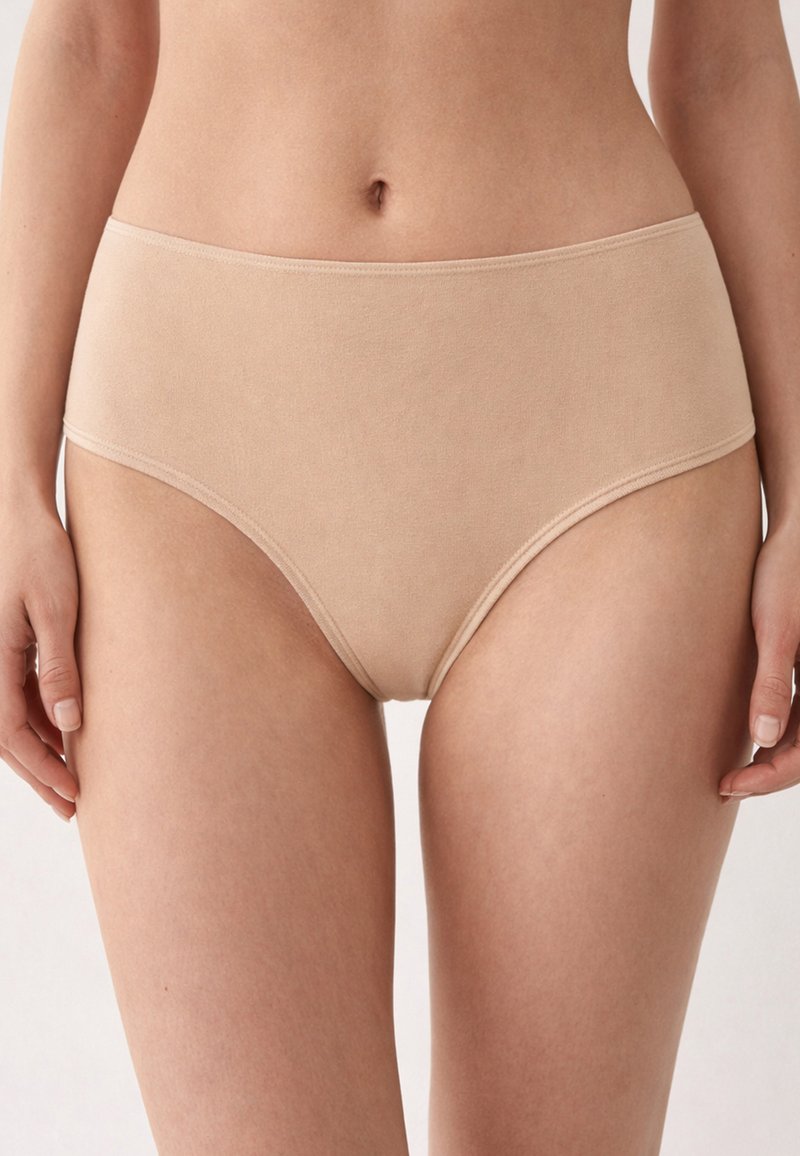 Donna che indossa biancheria intima in cotone beige a vita alta, in piedi con le mani rilassate ai lati, su uno sfondo bianco.