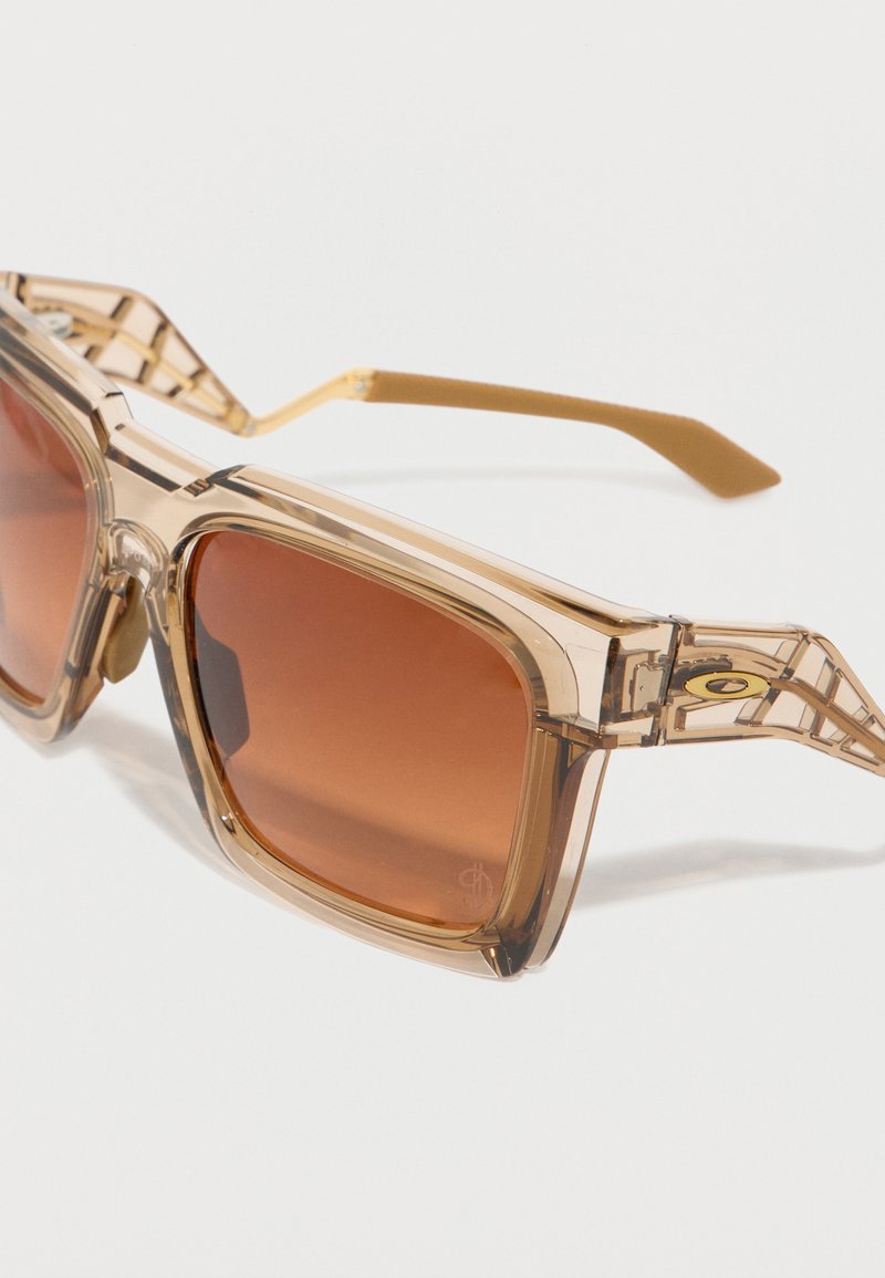 Lunettes de soleil à monture en plastique beige transparent avec des verres carrés teintés marron, posées en angle sur une surface blanche.