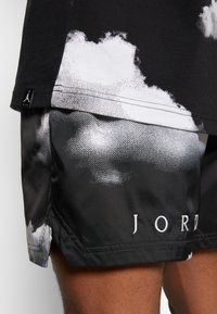 Detailní záběr osoby oblečené v černo-bílých šortkách Jordan s obláčkovými vzory a černém tričku se malou nášivkou loga Nike Jumpman.