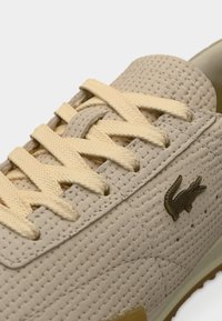 Baskets beiges texturées avec une tige tissée, des lacets jaunes et un petit logo crocodile en métal sur le côté. Semelle en caoutchouc avec motif antidérapant.