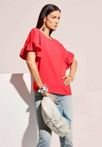 Rotes Oberteil mit Rü schenärmeln, kombiniert mit hellblauen Jeans. Das Modell hält eine weiche, hellfarbige Clutch und trägt silberne Accessoires.