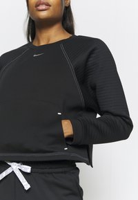 Donna che indossa un felpa nera Nike con maniche texture e pantaloni bianchi con logo Nike, con la mano nella tasca della felpa.