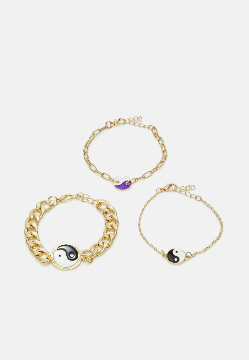 Três pulseiras douradas com pendentes de yin-yang. Uma tem uma corrente grossa, outra é uma corrente fina e delicada, e uma tem um pequeno encanto circular.
