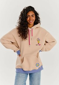 Béžová oversized mikina s ružovými sťahovacími šnúrkami, prednou kapsou a karikatúrou. Má svetlomodré manžety a nášivku s logom Looney Tunes.