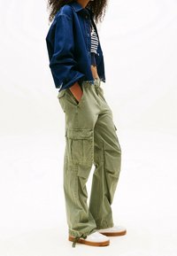 Syvänsininen farkkutakki, lyhennetty malli; oliivinvihreät cargo-housut, joissa on useita taskuja, rento istuvuus; yhdistetty valkoisiin lenkkareihin, joissa on beige pohja.