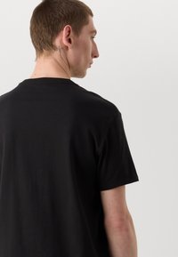 Svart bomulls T-skjorte med kort erm og rund halsutskjæring, med en glatt tekstur. Har et subtilt tatovering synlig på nakken.