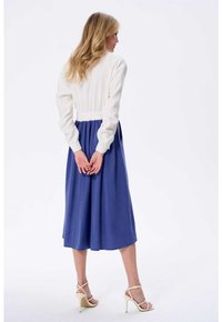 Długa niebieska sukienka z białą elastyczną talią i długimi rękawami. Tkanina wydaje się gładka, a sukienka sięga do połowy łydki. Beżowe sandały na obcasie.