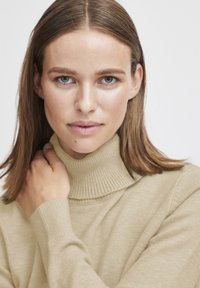 Beige polotröja gjord av mjukt stickat material, med ribbad krage och ärmslut, samt en slät yta och avslappnad passform.