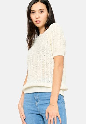 Maglione a maglia color crema con maniche corte, caratterizzato da un motivo testurizzato e orlo a costine, indossato sopra jeans azzurri chiari.