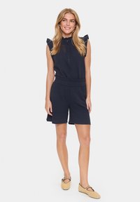 Marineblauer kurzer Jumpsuit mit kragenähnlichem Halsauschnitt, Knopfverschluss vorne, Rüschenärmeln, elastischem Bund und strukturiertem Stoff. Getragen mit beigen Ballerinas.