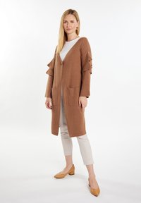 Bruine gebreide cardigan met uitlopende mouwen, sluiting met voorknoop, twee voorzakken en een middenlange snit, gecombineerd met beige broeken en tan hakken.
