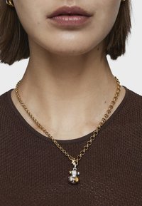 El collar de cadena dorada presenta un charm multicolor con acentos esféricos en plata, oro y bronce, usado sobre una blusa texturizada marrón.