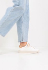 Λευκό δερμάτινο αθλητικό παπούτσι με μπεζ τόνους στη φτέρνα, με υφή στα κορδόνια και στρογγυλεμένη μύτη, συνδυασμένο με ανοιχτό μπλε τζιν παντελόνι με γυρισμένα μανίκια.