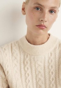 Maglione a maglia cable color crema con scollatura a girocollo a costine. Presenta motivi testurizzati e un tessuto in lana morbido. Inquadratura ravvicinata sulla parte superiore del corpo.