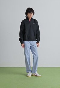 Personne avec des cheveux bouclés portant un sweat à capuche noir avec fermeture éclair à quart, un pantalon bleu clair et des baskets blanches, se tenant sur un sol vert contre un mur gris.