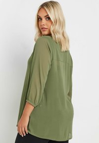 Blusa leggera verde oliva con maniche a tre quarti, polsi arricciati e orlo arrotondato; dettaglio della parte posteriore leggermente trasparente.