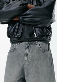 Schwarze Bomberjacke aus Kunstleder mit gerafften Ärmeln, getragen über einem grauen Sweatshirt. Graue Denim-Jeans mit geradem Schnitt und Vorder-Taschen.